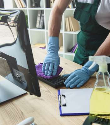 einigungskraft mit blauen Gummihandschuhen reinigt eine Computertastatur im Büro mit einem lila Tuch und Reinigungsmittel.