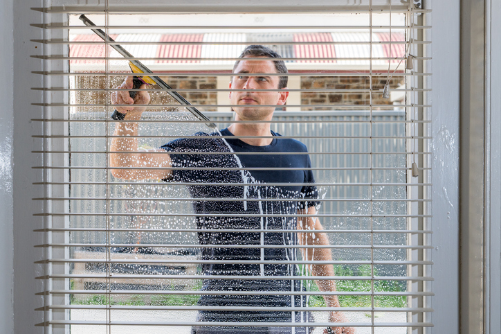 Professioneller Fensterputzer reinigt ein Fenster mit einem Fensterwischer, gesehen durch Jalousien.