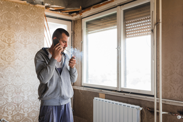 Mann in renovierungsbedürftigem Raum, der am Telefon spricht und eine Zigarette anzündet.