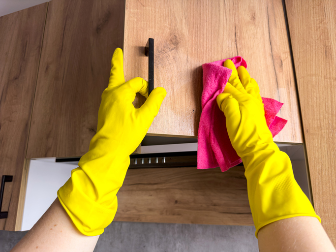 Gelbe Handschuhe reinigen eine Holzschranktür mit einem pinken Lappen.