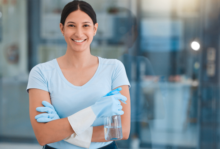 Lächelnde Reinigungskraft der Schlueter Group mit blauen Handschuhen und Sprühflasche in einem Büro. Im Hintergrund helle Büromöbel und Fenster.