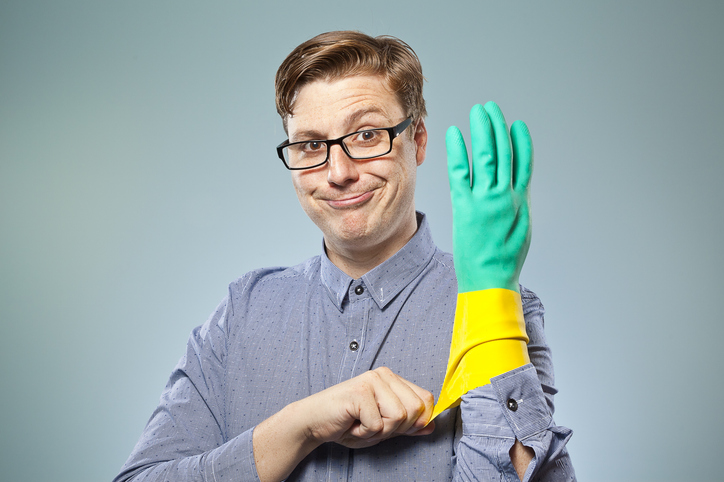 Ein Mann mit Brille und blauem Hemd zieht sich auf humorvolle Weise einen gelb-grünen Gummihandschuh über die rechte Hand. Er schaut dabei mit einem verschmitzten Lächeln in die Kamera.