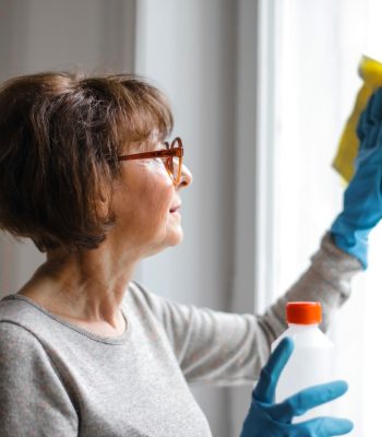 Reinigungskraft mit blauen Gummihandschuhen reinigt ein Fenster mit einem gelben Tuch und Reinigungsmittel.